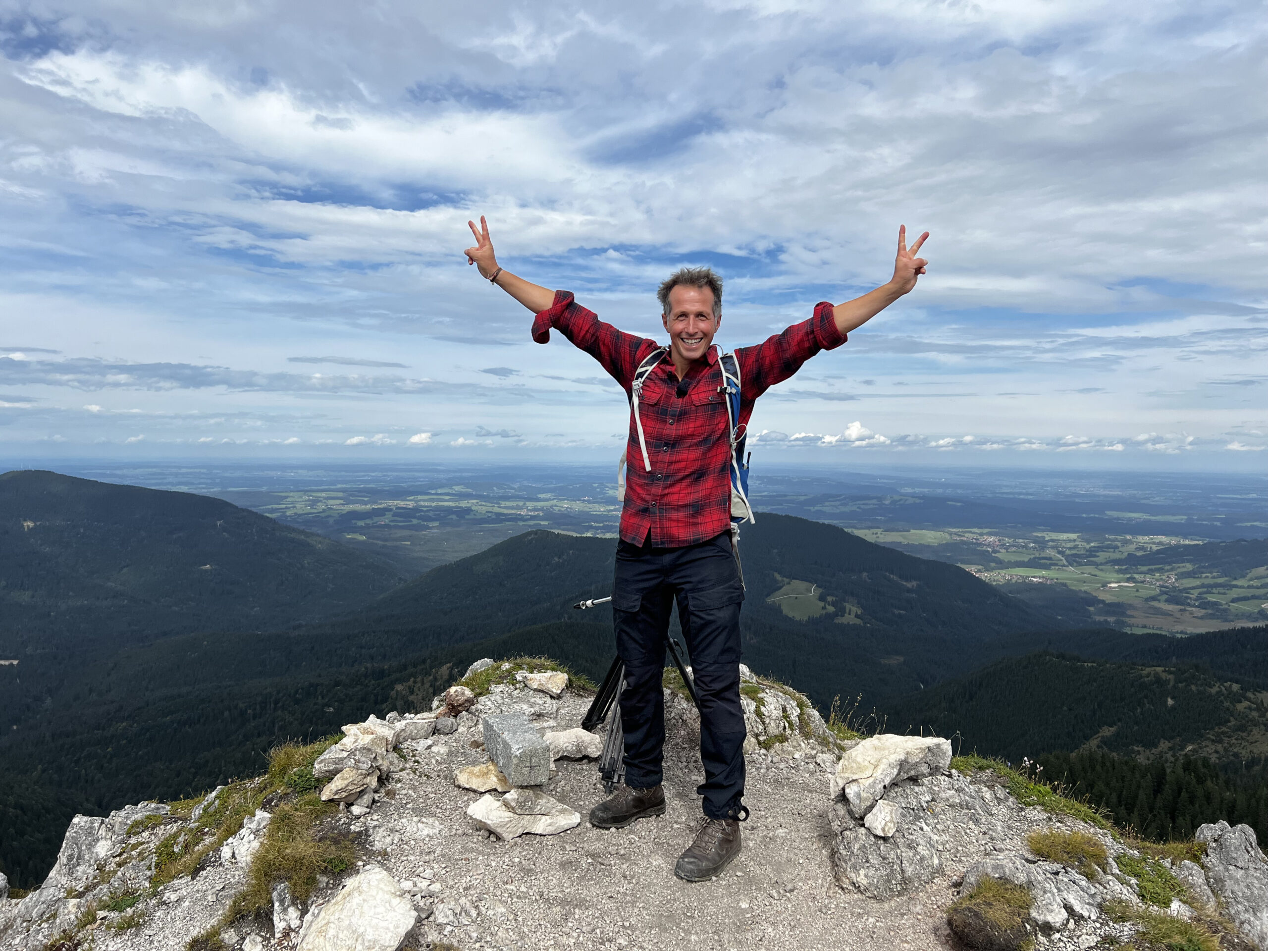 Ein Mann steht mit ausgestreckten Armen auf einem Berggipfel und formt mit den Fingern ein V. Im Hintergrund die umliegende Landschaft.